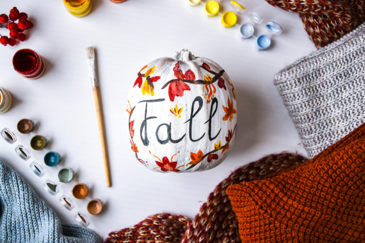 White pumpkin with painted leaves and the word fall on a table with paint, paint brushes, and fall sweaters