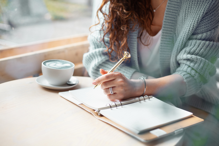 Young woman in blue warm sweater sitting near the big window of coffee shop and writing a Christmas shopping list with cup of blue latte. Planning Christmas holidays. Organising and planning concept.